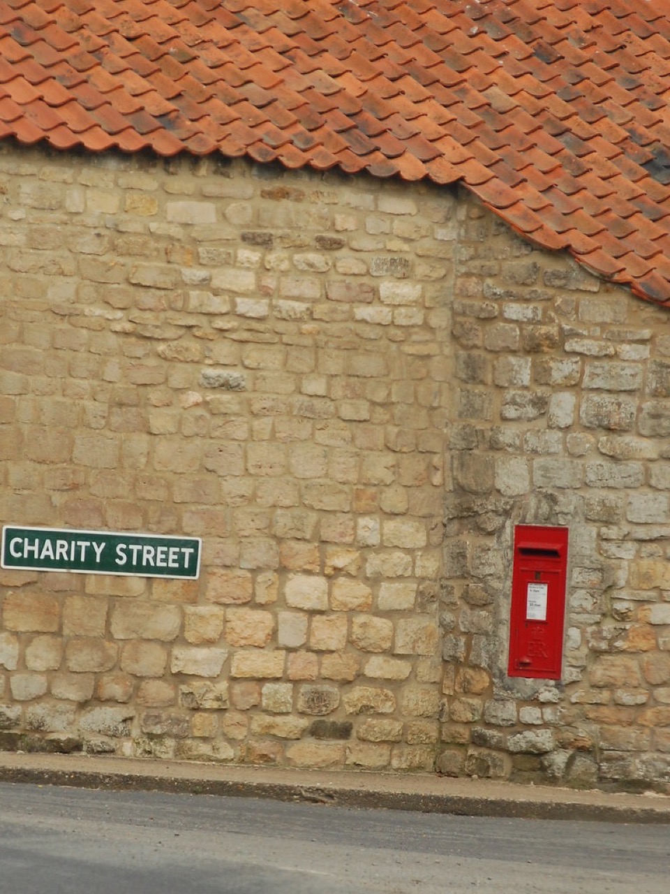 A limestone rubble wall under a red pantile roof with a street sign and a post box, at Carlton Scroop in Lincolnshire